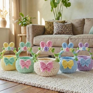 Può includere: Una fila di cinque cestini pasquali all'uncinetto colorati. Ogni cestino ha un fiocco, un manico con decorazioni floreali e dettagli di orecchie di coniglio. I colori includono giallo, verde menta, crema, azzurro e lavanda.