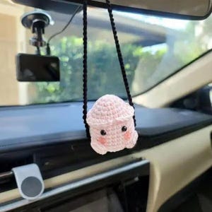 Könnte beinhalten: Ein rosa gehäkeltes Plüschtier mit einem lächelnden Gesicht hängt an einer schwarzen Schnur in einem Auto. Das Spielzeug ist am Rückspiegel befestigt.