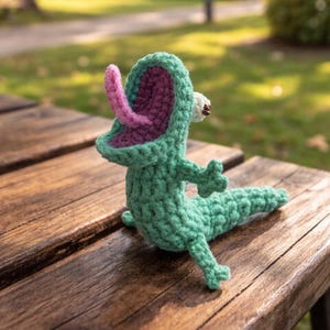 May include: A handmade crochet creature in shades of green, with a pink tongue and purple mouth. The whimsical design features small arms and legs, and is sitting on a wooden surface. The background is blurred, suggesting an outdoor setting.