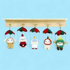 May include: Five crocheted stuffed toys hanging from a wooden shelf. Each toy is wearing a parachute and has a different design. The toys are a white bear wearing a red and white Santa suit, a blue penguin wearing a white and blue suit, a white cat wearing a red and white Santa hat, a brown dog wearing a green and brown Christmas tree costume, and a white bunny wearing a brown and white costume.