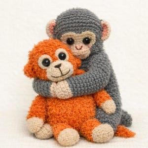 以下が含まれることがあります： 2匹のかぎ針編みの猿のぬいぐるみ。1匹はグレーで、もう1匹（オレンジ色）を抱きしめています。どちらも黒い目、タン色の顔、ベージュ色の手足です。グレーの猿はピンクの耳で、オレンジの猿はふわふわした質感です。