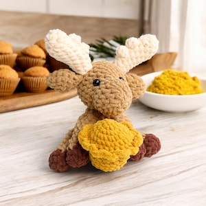 May include: A brown and white crocheted moose holding a yellow crocheted pie. The moose has large, floppy ears and a sweet expression.