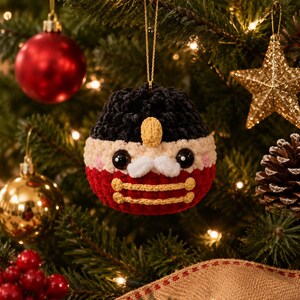 Peut inclure: Une d&eacute;coration de No&euml;l en crochet en forme de casse-noisette rond, avec un chapeau noir, un visage beige aux yeux en boutons noirs, une moustache blanche et un corps rouge orn&eacute; de deux lignes horizontales en fil dor&eacute;.