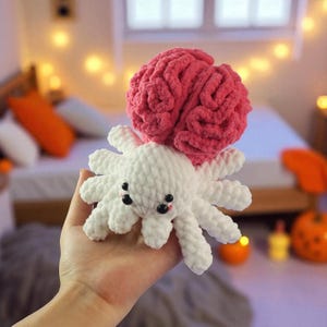 May include: A handmade crochet octopus with a pink brain on top. The octopus is white with black eyes and a small pink mouth. The brain is a vibrant pink color. The toy is soft and plush.
