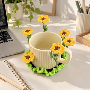 May include: A cream-coloured ceramic mug with vertical ridges, resting on a circular green crocheted coaster. The coaster features five attached crocheted sunflowers with yellow petals and brown centres that extend upwards around the mug.