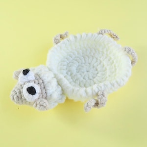 May include: A handmade crochet sheep-shaped basket in white and beige. The basket has a fluffy, textured body and a detailed head with black eyes. The sheep has beige legs and ears, set against a yellow background.