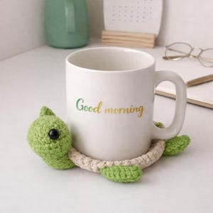 May include: A white ceramic mug with the words "Good morning" in green and yellow script. The mug is cradled by a green and beige crocheted turtle coaster. The turtle coaster has a green head and feet, with a beige body.
