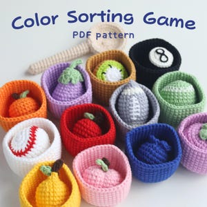 以下が含まれることがあります： 色とりどりの、かぎ針編みの玩具の果物とスポーツボールが、それぞれ対応するバスケットに入っています。野球ボール、アメフトボール、黒い8ボールが含まれています。「Color Sorting Game PDF pattern」の文字が上部にあります。