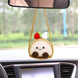 May include: A crocheted plush toy shaped like a roll cake with a smiling face, a red strawberry, and white whipped cream on top. The toy is hanging from a rearview mirror in a car.