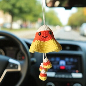 Op de afbeelding: Een handgemaakte, gehaakte candy corn auto-hanger. De hanger is geel, oranje en wit, met een lachend gezicht en bungelende candy corn accenten. Hij hangt aan een wit touwtje en voegt een vleugje vrolijkheid toe aan het interieur van een auto.