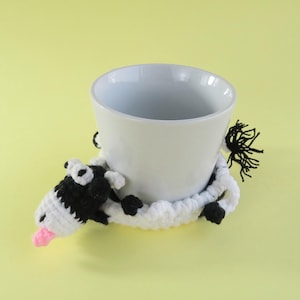 May include: A white ceramic mug rests on a handmade crochet coaster shaped like a cow. The cow coaster features black and white details, including a pink tongue, and a fluffy black tail. The background is a solid light yellow.