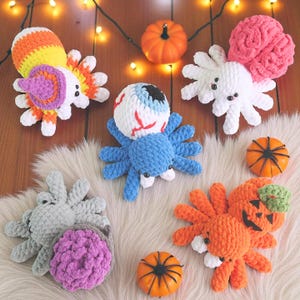 May include: A selection of handmade crochet spiders with a Halloween theme. The spiders are in various colours, including blue, orange, and white, with designs such as an eyeball, a jack-o'-lantern, and a brain. Small orange pumpkins are also visible.