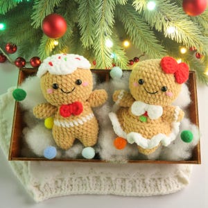 May include: Two crocheted gingerbread figures, one with a red bow tie and the other with a red bow, sit in a wooden tray with faux snow. The gingerbread people have smiling faces and are surrounded by Christmas decorations.