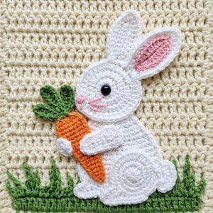 May include: A crocheted white bunny holding an orange carrot with green leaves, set against a cream-colored background. The bunny has pink inner ears and black eyes. Green crocheted grass is at the bottom.
