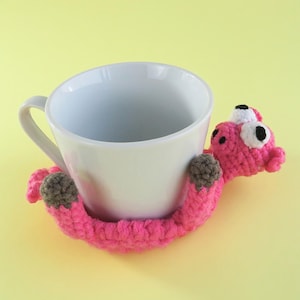May include: A white ceramic mug is cradled by a handmade, pink crocheted creature. The creature has brown paws and white and black eyes. The mug and creature are set against a yellow background.