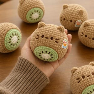 Può includere: Peluche a forma di gatto-kiwi all'uncinetto. I giocattoli sono marroni con fette di kiwi verdi e bianche sul fondo. Ogni peluche ha una faccia da gatto con piccole orecchie e baffi. Uno è tenuto in mano, altri sono sparsi su una superficie di legno.