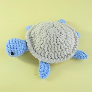 May include: A handmade crochet turtle plushie. The turtle has a light gray shell, a blue head, and four blue flippers. The turtle's shell is round and textured, and the head has a small black eye. The plushie is on a yellow background.