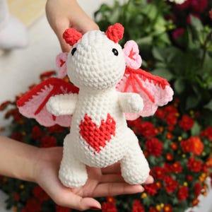 May include: A handmade crochet dragon toy in white with red and pink accents. The dragon has a red heart on its chest, red horns, and pink and red wings. The toy is held in a person's hands against a background of red flowers.