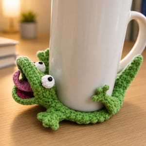 May include: A handmade, green crochet alligator mug hugger with white eyes and a pink tongue. The alligator is wrapped around a white mug. The mug hugger is designed to keep your drink warm and add a touch of whimsy to your morning routine.