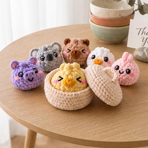 Puede incluir: Una colección de pequeñas figuras de peluche tejidas a ganchillo, que incluye un cerdo morado, un koala gris, un oso marrón, un pájaro blanco, un conejo rosa y un pollito amarillo dentro de una cesta de ganchillo beige con tapa.