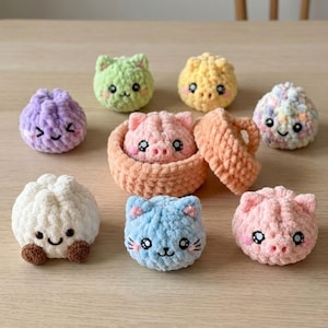 Könnte beinhalten: Eine Sammlung kleiner, runder, gehäkelter Plüschfiguren in verschiedenen Farben wie Lila, Grün, Gelb, Weiß, Blau und Rosa. Einige ähneln Tieren wie Schweinen und Katzen, andere sind runde Formen. Ein rosa Schweinchen sitzt in einem pfirsichfarbenen Korb.