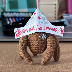 Puede incluir: Una criatura de ganchillo marrón con cuatro patas y un cuerpo. Lleva un sombrero de papel con las palabras "SAVIOR OF THE UNIVERSE" en rojo. Una estrella plateada está unida al cuerpo de la criatura.