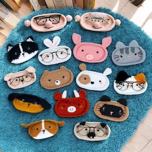 May include: A collection of 14 crocheted animal-shaped eyeglass holders. Each holder features a different animal face, including a pig, a cat, a dog, a bunny, a bear, and a bull. The holders are all made in different colors, including pink, gray, brown, white, and red.