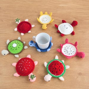 Puede incluir: Una colección de ocho posavasos de ganchillo con forma de tortuga dispuestos alrededor de una taza de cerámica blanca central. Los posavasos presentan varios diseños inspirados en frutas como sandía, kiwi y pitaya en rojo, verde, amarillo y blanco.