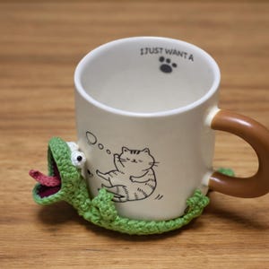 Puede incluir: Una taza de cer&aacute;mica blanca con un asa marr&oacute;n y el texto "I JUST WANTA". La taza presenta una ilustraci&oacute;n de gato de dibujos animados y est&aacute; rodeada por un posavasos de lagarto de ganchillo verde con una lengua rosa.