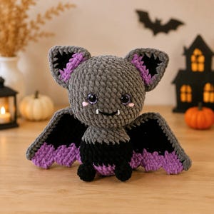 May include: A crocheted bat plush toy featuring a gray head and body, black wings with purple scalloped edges, and large purple-tinted eyes. The bat has small white fangs, pink cheek accents, and large ears with black and purple inner details.