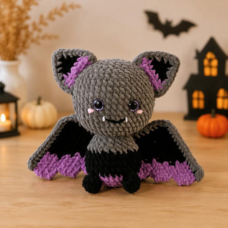 May include: A crocheted bat plush toy featuring a gray head and body, black wings with purple scalloped edges, and large purple-tinted eyes. The bat has small white fangs, pink cheek accents, and large ears with black and purple inner details.