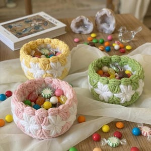 May include: Three handmade crochet baskets in yellow, pink, and green, each filled with colorful candies. The baskets have a white floral pattern. The image also includes a book, decorative stones, and scattered candies on a wooden surface.