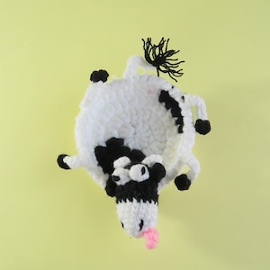 May include: A handmade crochet cow toy in white and black, with a pink tongue sticking out. The cow has black spots, hooves, and a tuft of black yarn for a tail. The toy is set against a light yellow background.