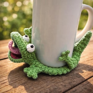 May include: A white ceramic mug held by a green crocheted alligator coaster. The alligator has white eyes, a pink tongue, and red mouth. The coaster is on a wooden surface.