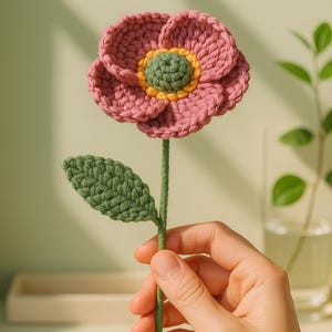 May include: A handcrafted crochet flower with a pink bloom, green centre, and yellow accents. The flower has a green stem and leaf. The craft item is held in a hand against a soft green backdrop.