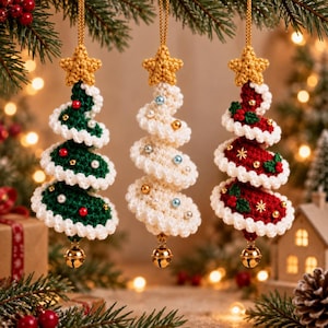 Op de afbeelding: Drie spiraalvormige kerstboomhangers van haakwerk die verticaal hangen. Elke boom is bekroond met een gouden ster, heeft witte geschulpte randen en is versierd met kleine kralen en een gouden belletje aan de onderkant in groen, wit en rood.