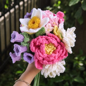 May include: A colorful crochet bouquet featuring a variety of flowers. The bouquet includes a large pink peony, white and yellow daffodils, and purple bell-shaped flowers. The flowers are made of yarn in shades of pink, white, yellow, green, and purple.