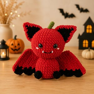 May include: A plush crochet bat toy featuring a round red body, large red and black ears, and wide red wings with black scalloped edges. The creature has two black plastic eyes, a small green stem on top, and two white fangs on its face.