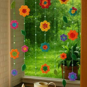 Op de afbeelding: Een kleurrijk gehaakt bloemengordijn met bloemen in verschillende maten in tinten oranje, geel, blauw, roze en paars. De bloemen zijn geregen op groen garen met kleine kralen en groene bladeren. Een decoratieve gieter staat eronder.