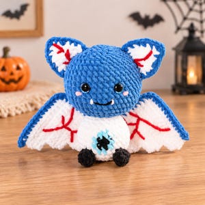 May include: A crocheted plush bat with a blue head, white body, and white wings featuring red vein-like embroidery. The bat has large black eyes, two small white fangs, and a single large blue eye centered on its white belly with black feet.