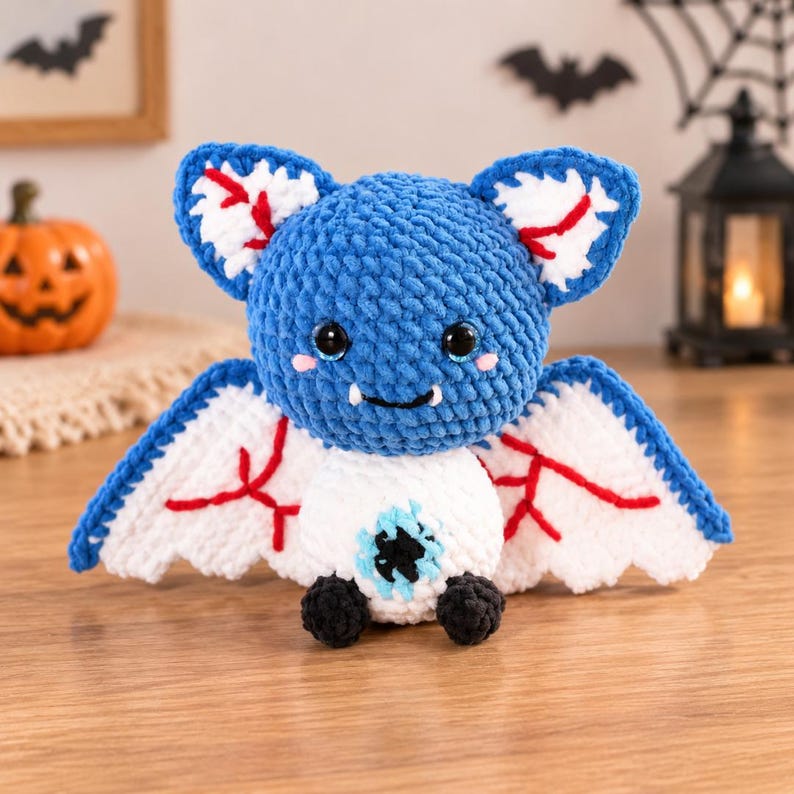 May include: A crocheted plush bat with a blue head, white body, and white wings featuring red vein-like embroidery. The bat has large black eyes, two small white fangs, and a single large blue eye centered on its white belly with black feet.