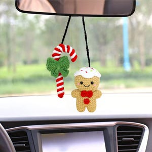 May include: A crocheted gingerbread man and candy cane hanging from a rear view mirror in a car. The gingerbread man is brown with white icing and sprinkles, and has a red bow tie. The candy cane is red and white with a green bow.