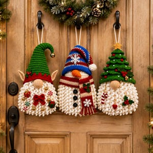 May include: Three hanging crochet ornaments featuring a green elf with a red bow, a snowman with a blue striped hat and scarf, and a gnome wearing a green Christmas tree hat. Each figure has a textured white beard and festive holiday accents.