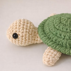 May include: A crocheted turtle toy with a cream-colored head and limbs, a green shell, and a black eye. The handmade turtle is a soft, plush toy.
