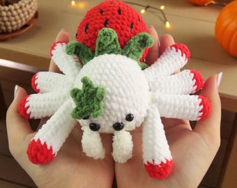 May include: A handmade crochet spider with white body and legs, red tips, and black eyes. A red strawberry with green leaves sits on top. The spider is held in open hands, with pumpkins and gourds in the background.