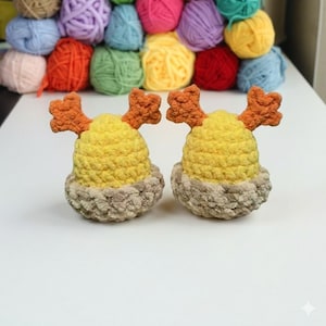 May include: Two handmade crochet crab hats. Each hat features a yellow body, a tan base, and orange crab claw accents. The hats are displayed on a white surface with a backdrop of colorful yarn balls.