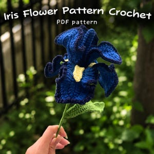 May include: A handmade crochet iris flower in shades of blue, with a yellow center and green stem and leaves. The text "Iris Flower Pattern Crochet" and "PDF pattern" are displayed at the top. The flower is held in a person's hand.