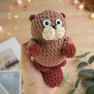 May include: A handmade crochet beaver plush toy. The beaver is primarily brown with lighter beige accents on the teeth and muzzle. It has large, googly eyes and small, dark red paws and tail. The toy is held in a hand.