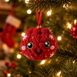 Peut inclure: Une d&eacute;coration ronde en crochet rouge avec une surface textur&eacute;e, de grands yeux en plastique noir et vert, un petit sourire brod&eacute; en noir et des joues rose clair. L'ornement est orn&eacute; de flocons de neige blancs brod&eacute;s et suspendu par un ruban rouge.