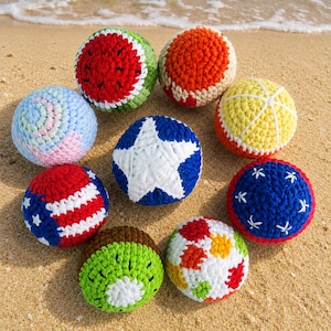 Puede incluir: Una colección de bolas de ganchillo coloridas, cada una con un diseño único. Las bolas presentan patrones como una sandía, una estrella y un kiwi. Las bolas se exhiben en una playa de arena, mostrando la artesanía hecha a mano y los colores vibrantes.